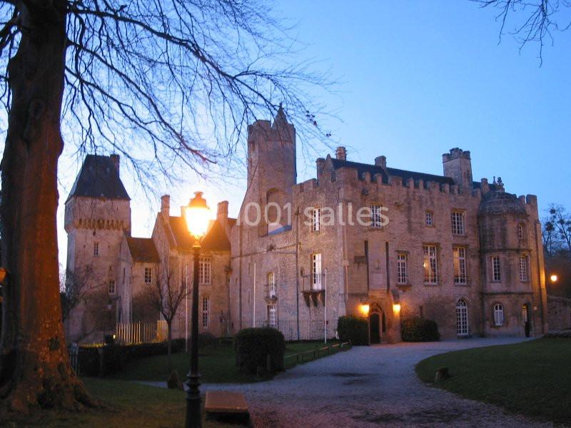 Château en pierre éclairé par des lampadaires au crépuscule, entouré d'arbres et d'une allée gravillonnée.