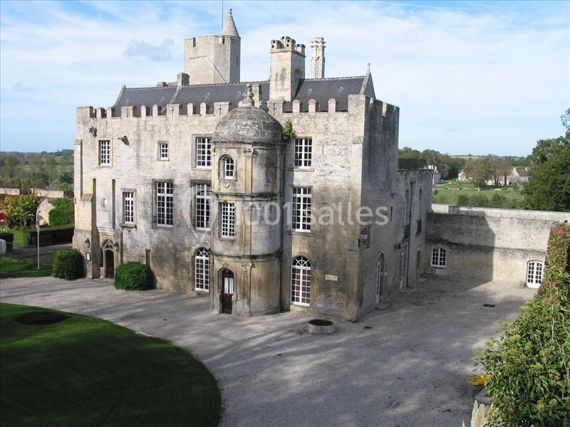 Château en pierre avec tourelles, fenêtres à meneaux et cour gravillonnée, entouré de pelouses et d'arbres.