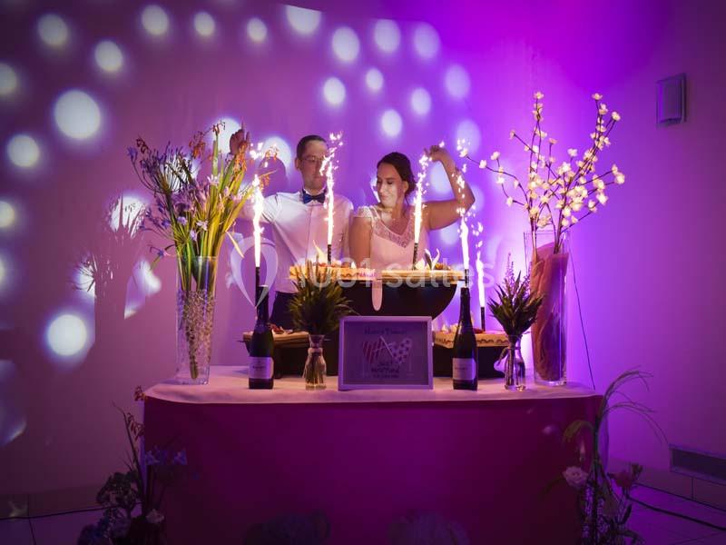 Un couple coupe un gâteau de mariage sur une table décorée, éclairée par des lumières violettes et des étincelles.