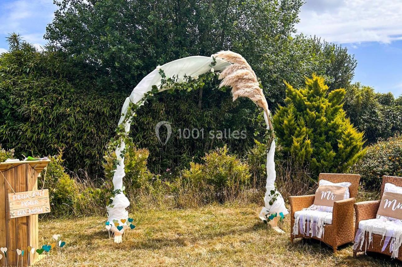 Arche de mariage décorée de tissu blanc, feuillage et pampas, entourée de chaises et végétation en extérieur.