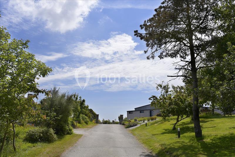 Route bordée de verdure menant à des maisons modernes sous un ciel partiellement nuageux.