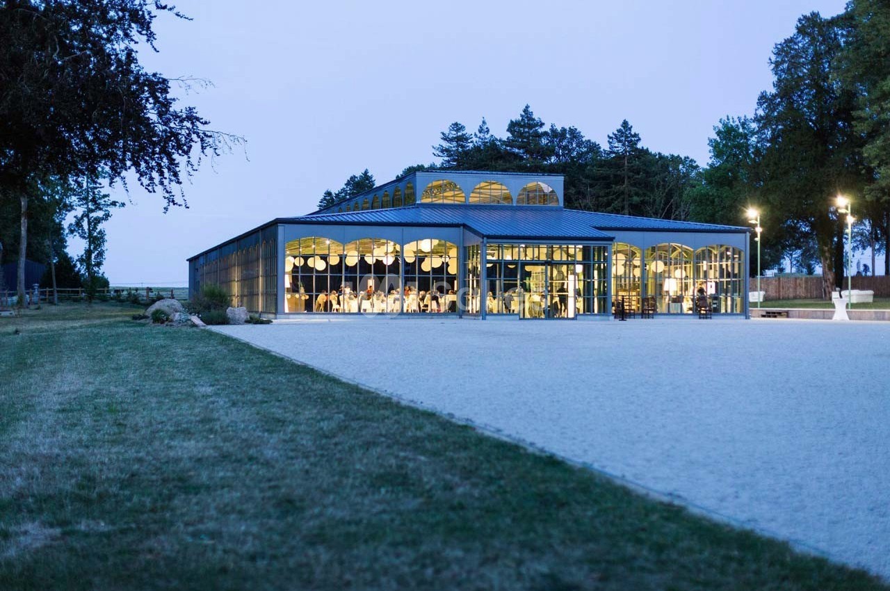 Bâtiment moderne avec de grandes baies vitrées illuminées, entouré d'arbres et d'une allée gravillonnée.