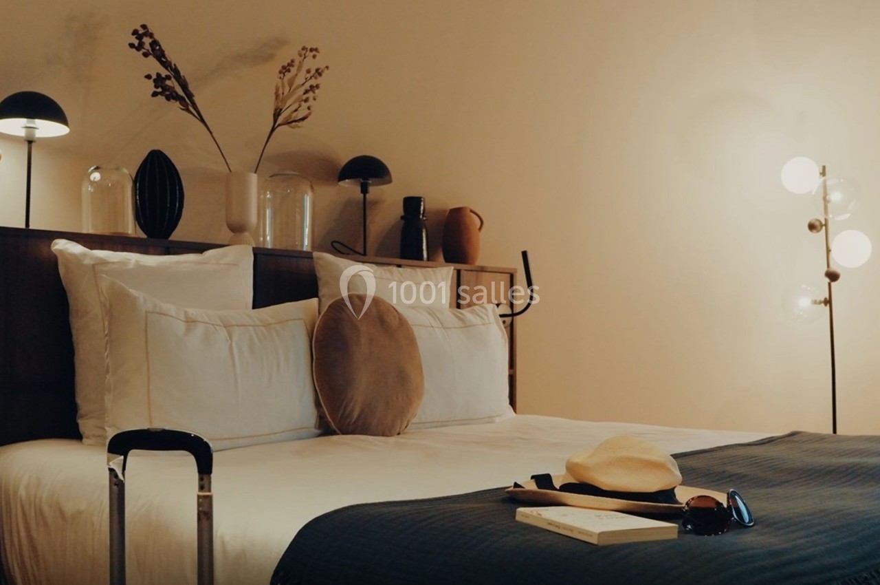 Lit double avec coussins et plaid, valise au pied, lampe allumée et décoration minimaliste sur une étagère en bois.