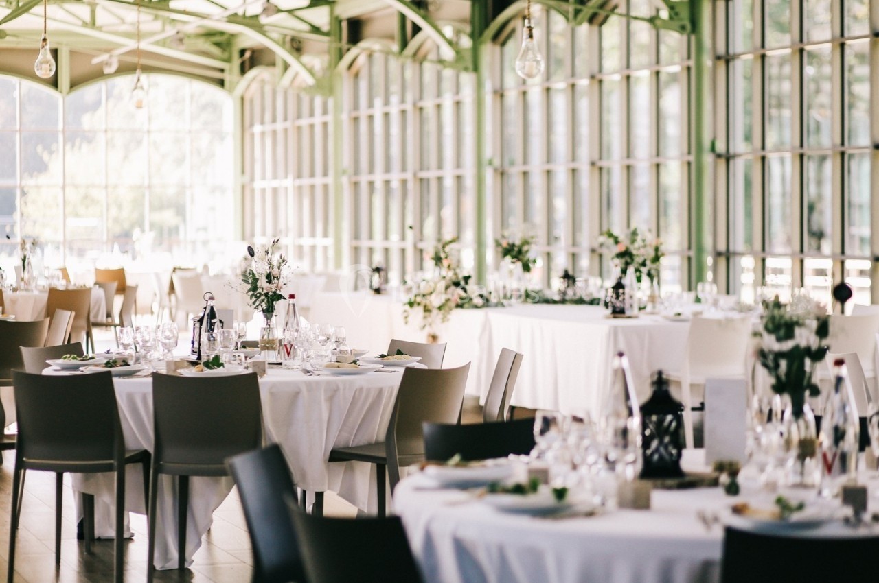 Salle lumineuse avec de grandes baies vitrées, tables rondes dressées avec nappes blanches et décorations florales.