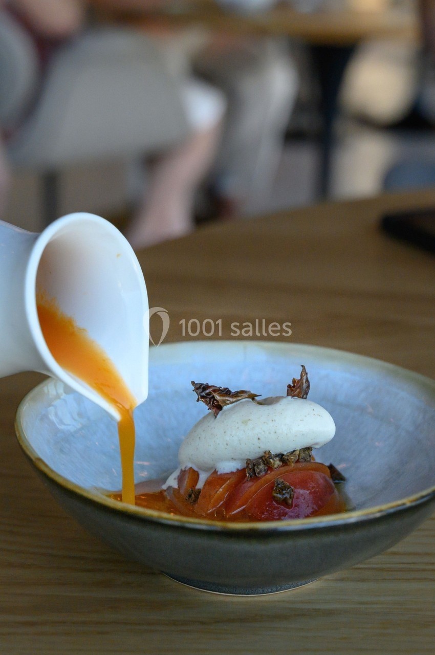 Un coulis orange est versé dans une assiette contenant des tomates, une mousse blanche et des garnitures croustillantes.