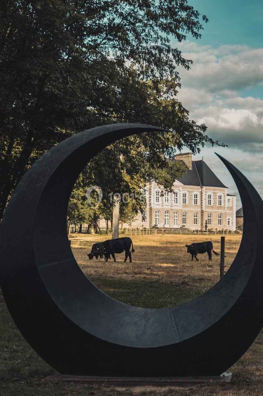 Sculpture en forme de croissant encadrant des vaches paissant dans un champ devant un manoir sous un ciel partiellement…