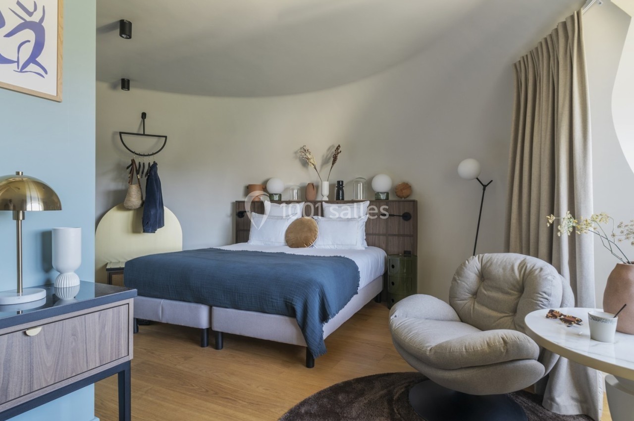 Chambre moderne avec lit double, fauteuil, table basse, décor minimaliste et lumière naturelle entrant par une fenêtre.