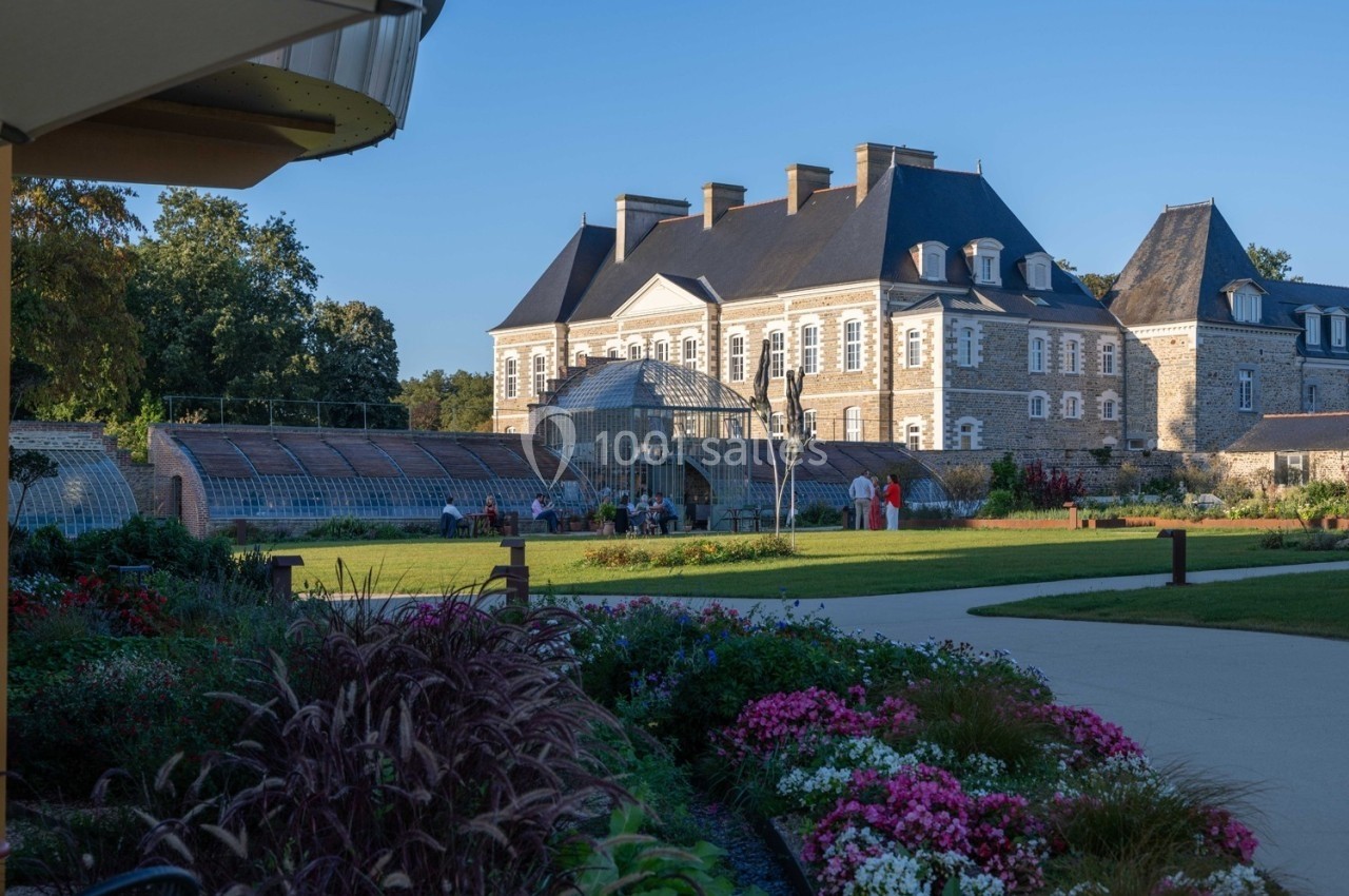 Vue d'un château en pierre entouré de jardins fleuris et d'une serre, par une journée ensoleillée.