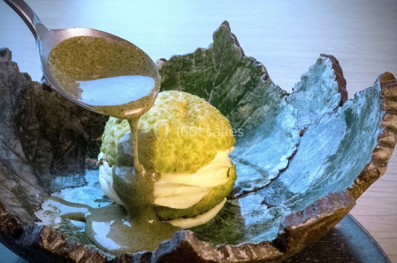 Un dessert avec une pâte verte, garni de crème, est nappé de sauce verte dans une assiette en céramique texturée.