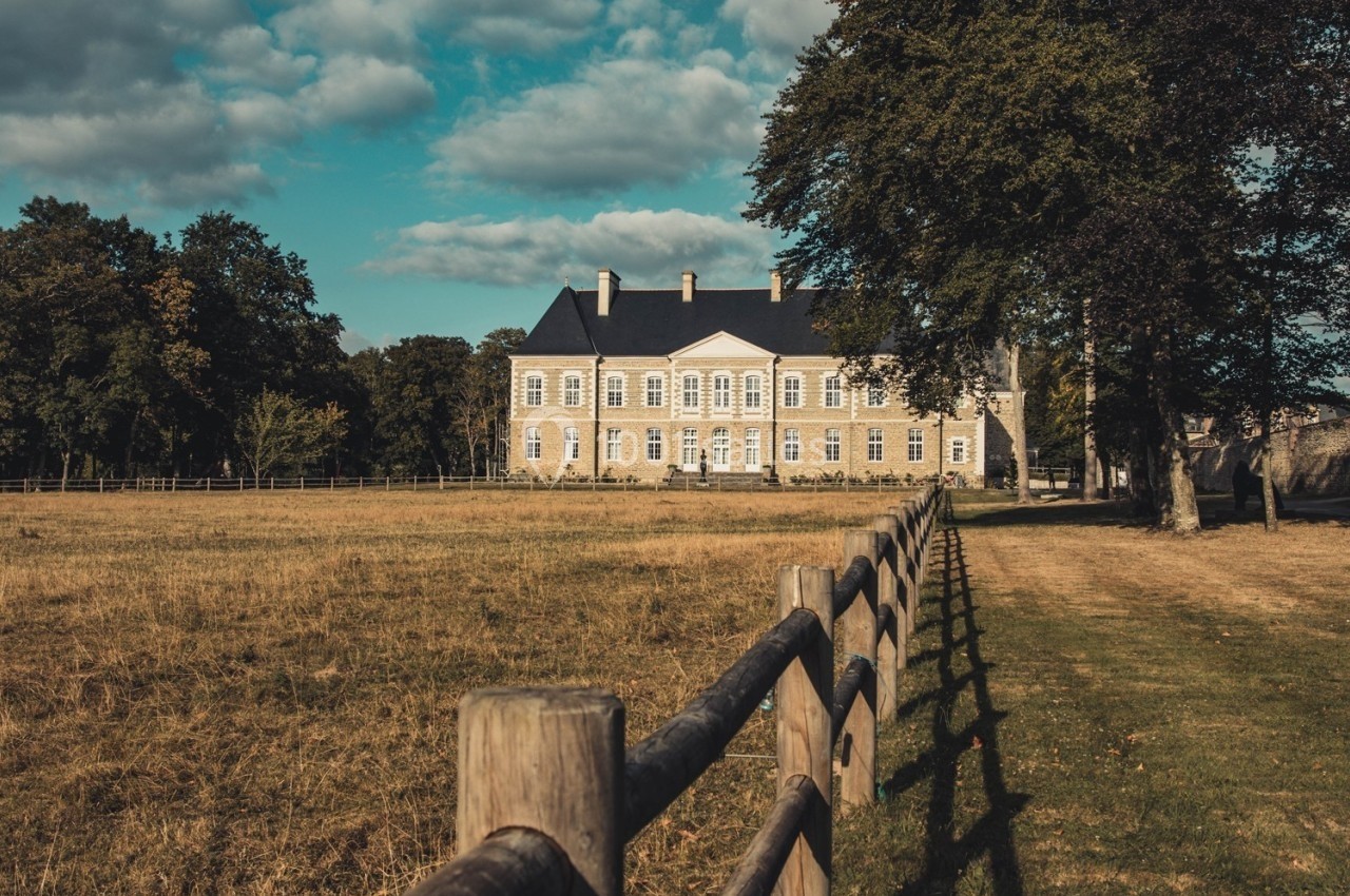 Manoir en pierre entouré de champs et d'arbres, avec une clôture en bois au premier plan sous un ciel partiellement nuageux.