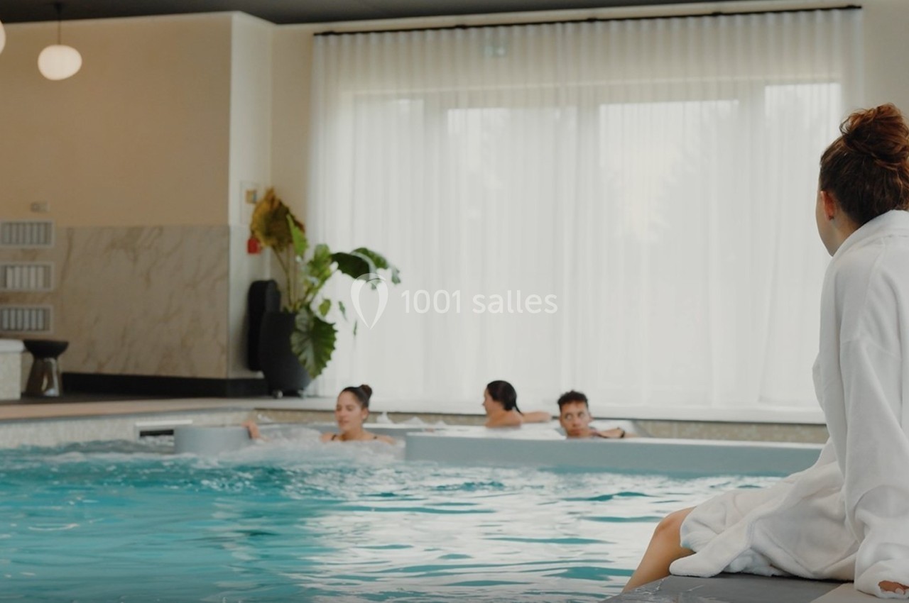 Une femme en peignoir assise au bord d'une piscine intérieure, observant des personnes se baignant.