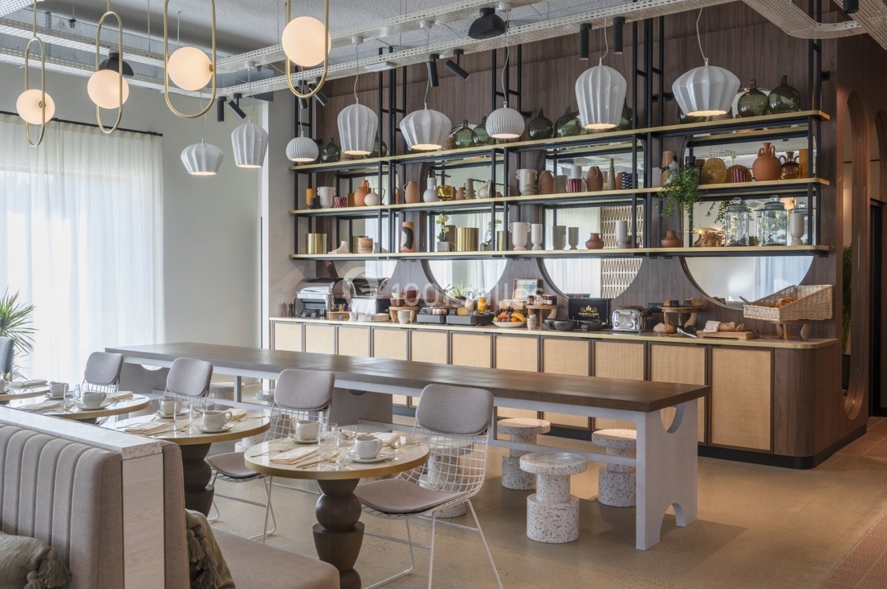 Salle de restaurant lumineuse avec tables dressées, longue table en bois et étagères décorées d'objets et vaisselle.
