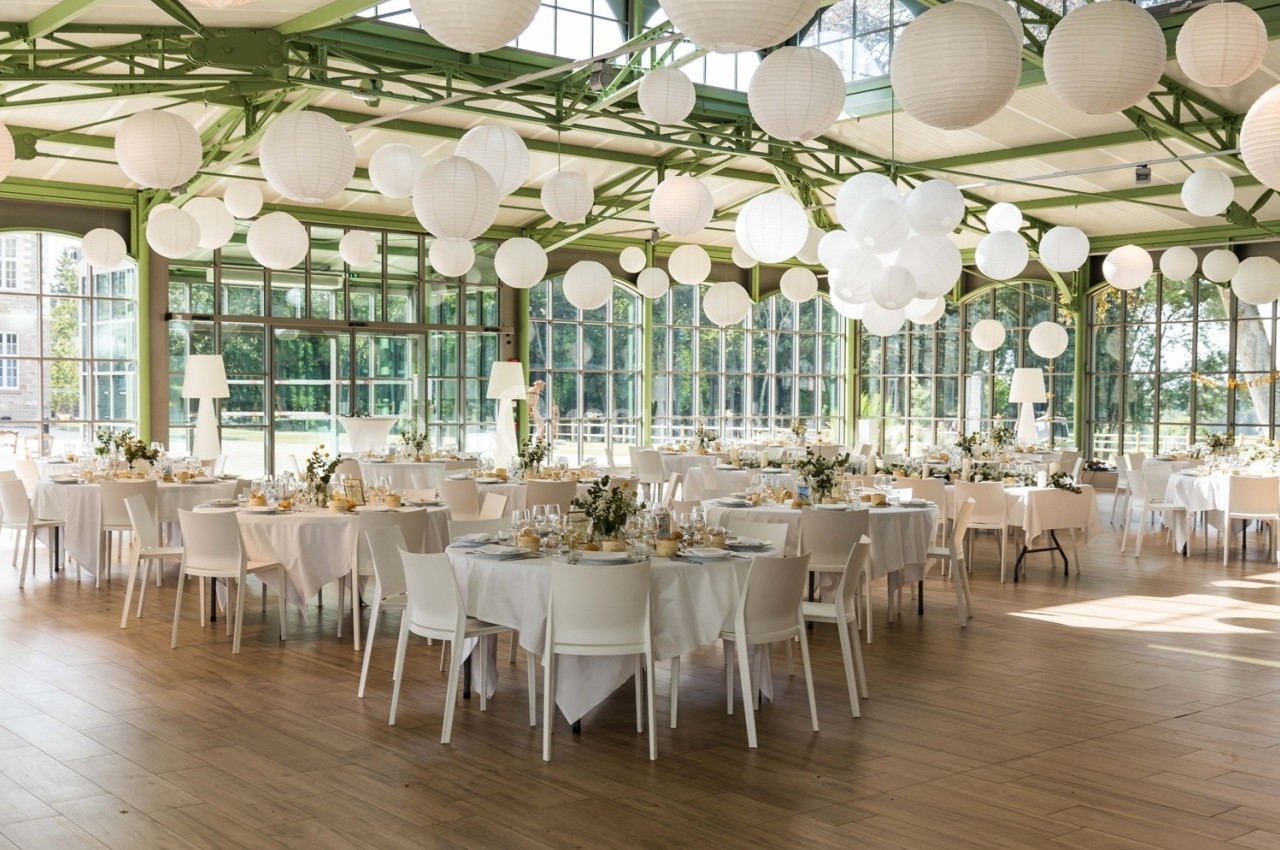 Salle de réception lumineuse avec tables rondes dressées, décorée de lanternes blanches suspendues.