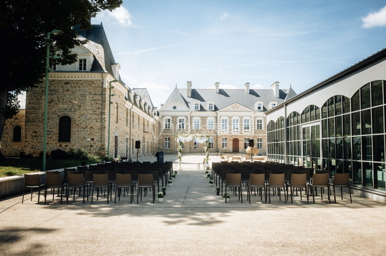 Chaises alignées en extérieur devant un château en pierre, préparées pour une cérémonie sous un ciel ensoleillé.