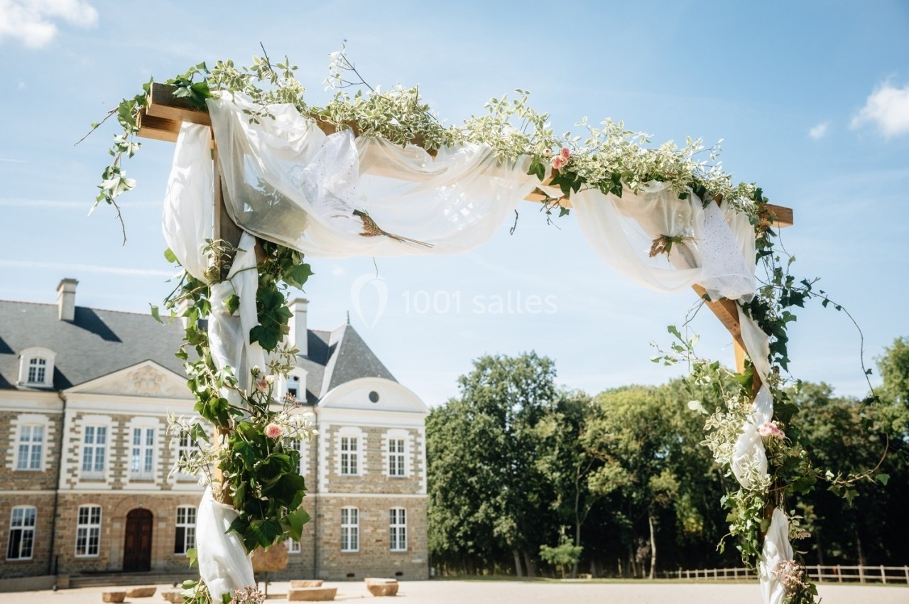Arche de mariage décorée de fleurs et de voilages blancs, devant un bâtiment ancien sous un ciel bleu.