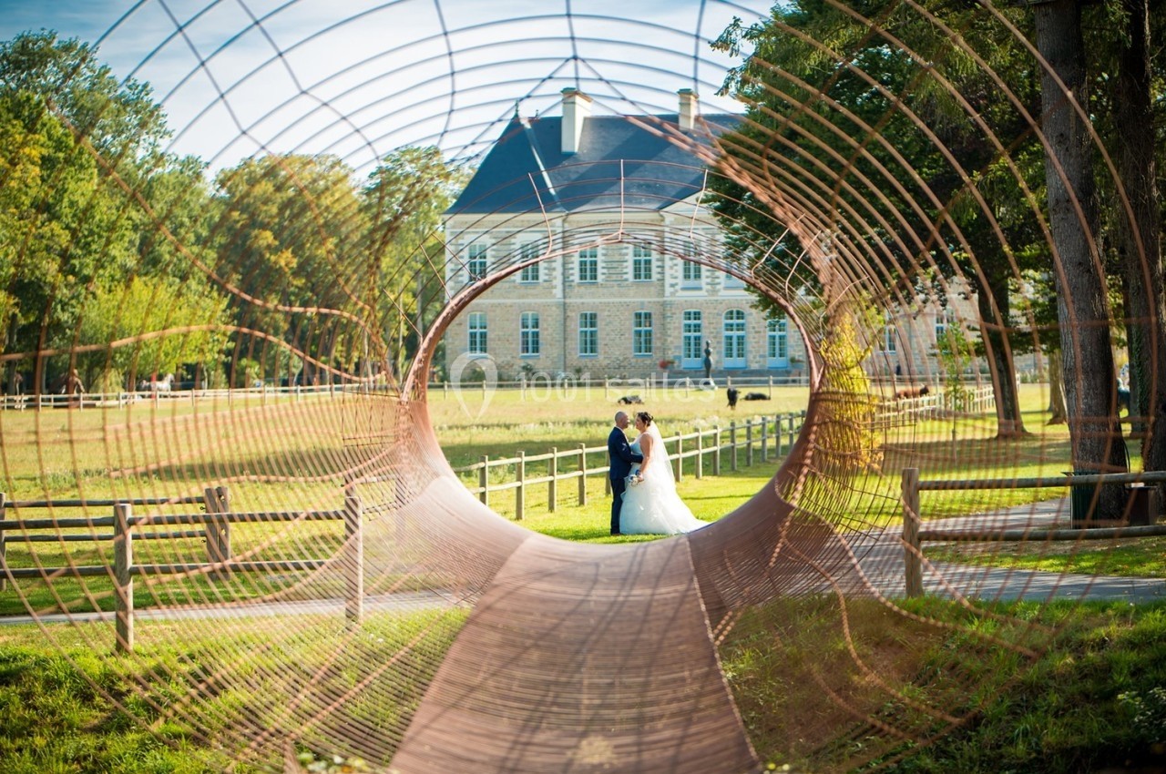 Un couple de mariés pose dans un parc, encadré par une structure circulaire en métal, avec un manoir en arrière-plan.