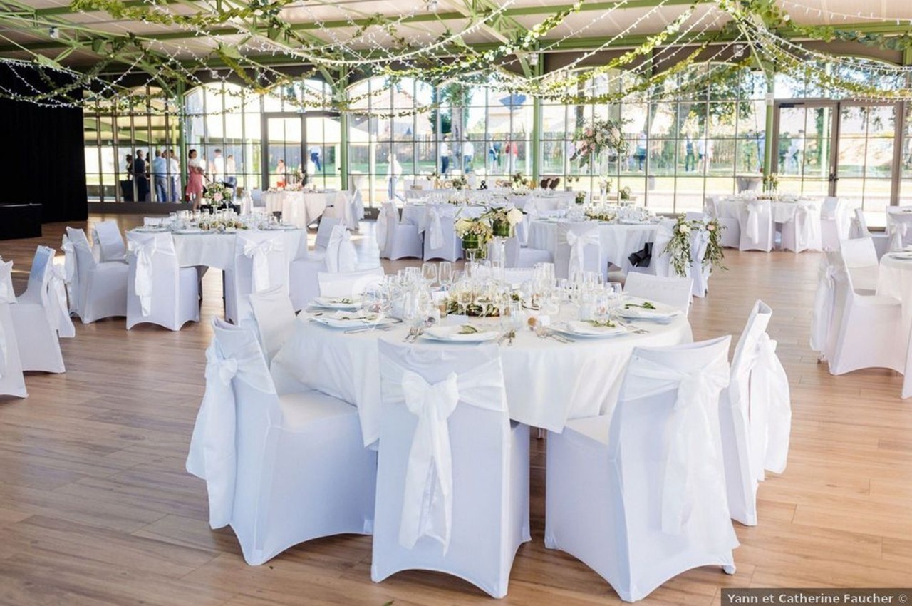 Salle de réception lumineuse décorée de guirlandes et de tables rondes dressées avec nappes blanches et fleurs.