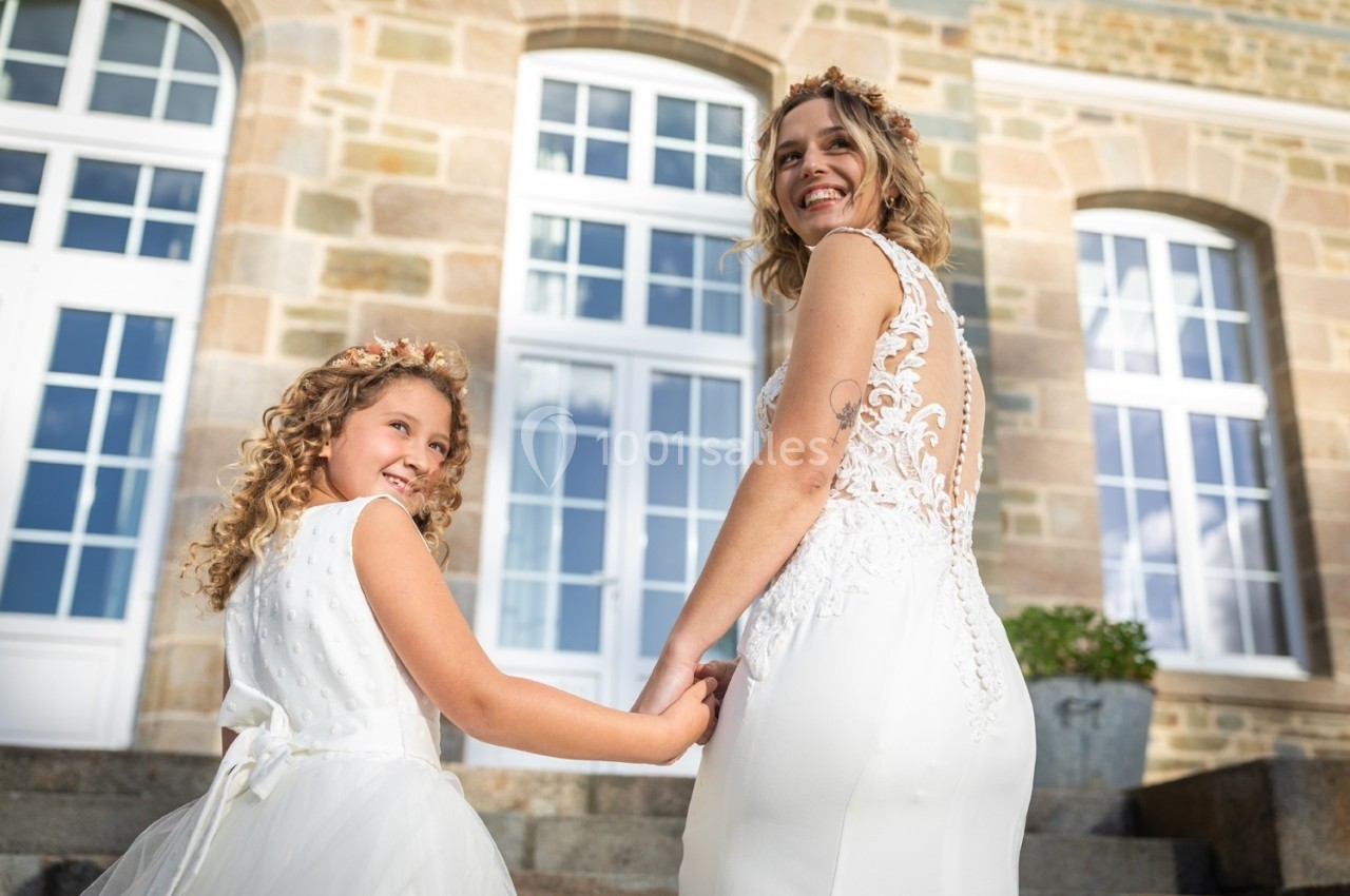Une mariée souriante tenant la main d'une jeune fille devant un bâtiment en pierre avec de grandes fenêtres.