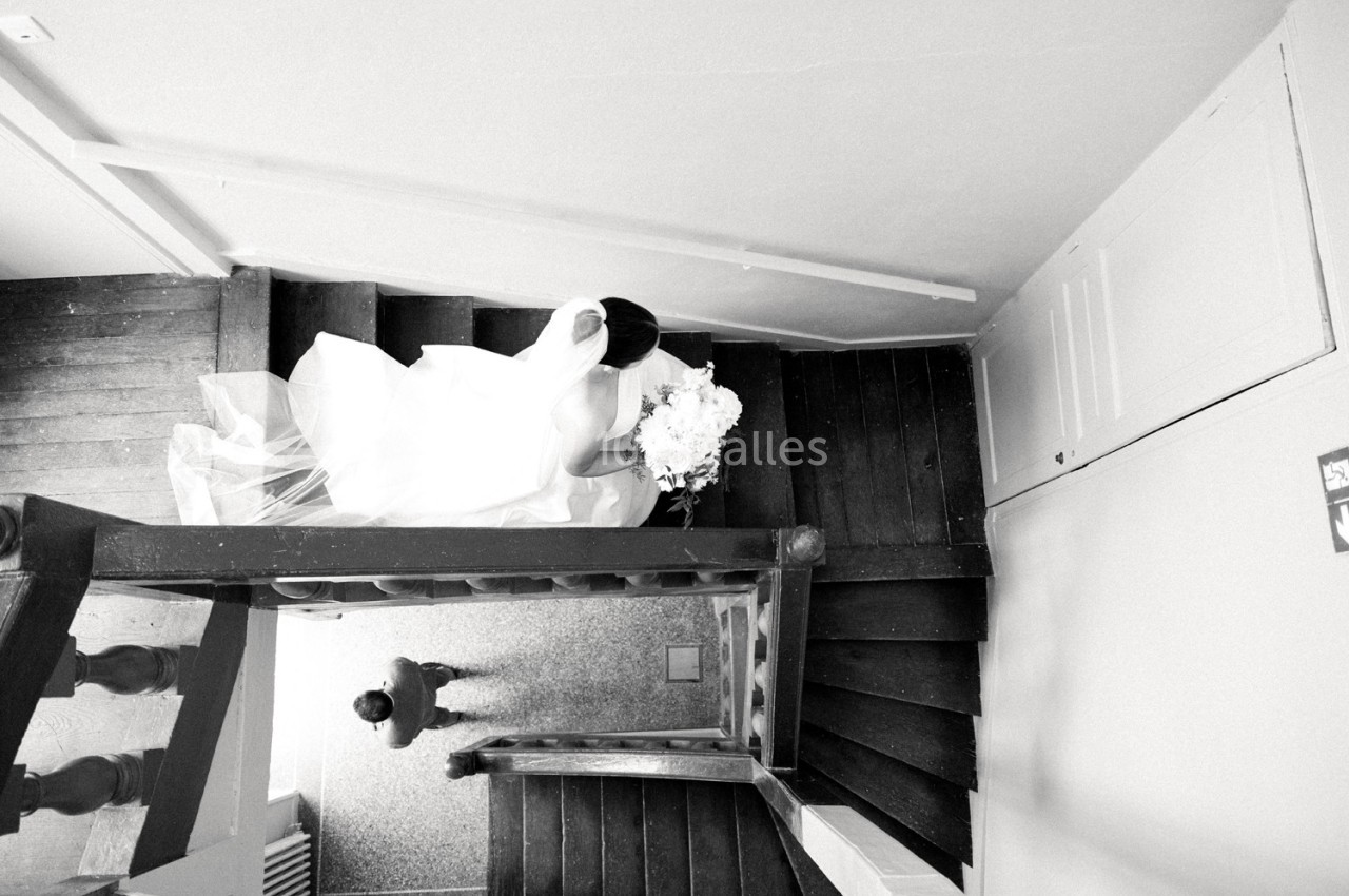 Une mariée en robe blanche descend un escalier en bois, vue de dessus, tenant un bouquet de fleurs.