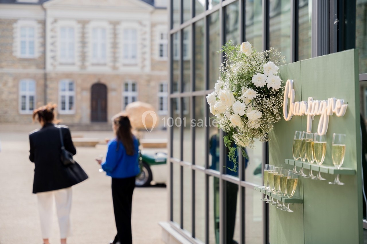 Mur décoratif avec des verres de champagne et des fleurs blanches, deux personnes discutant devant un bâtiment élégant.