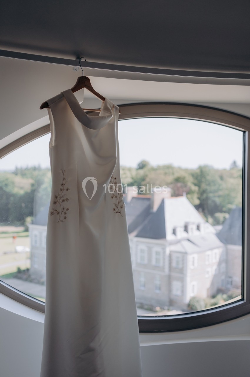 Robe blanche élégante suspendue à un cintre devant une fenêtre ovale avec vue sur un bâtiment historique et un paysage…