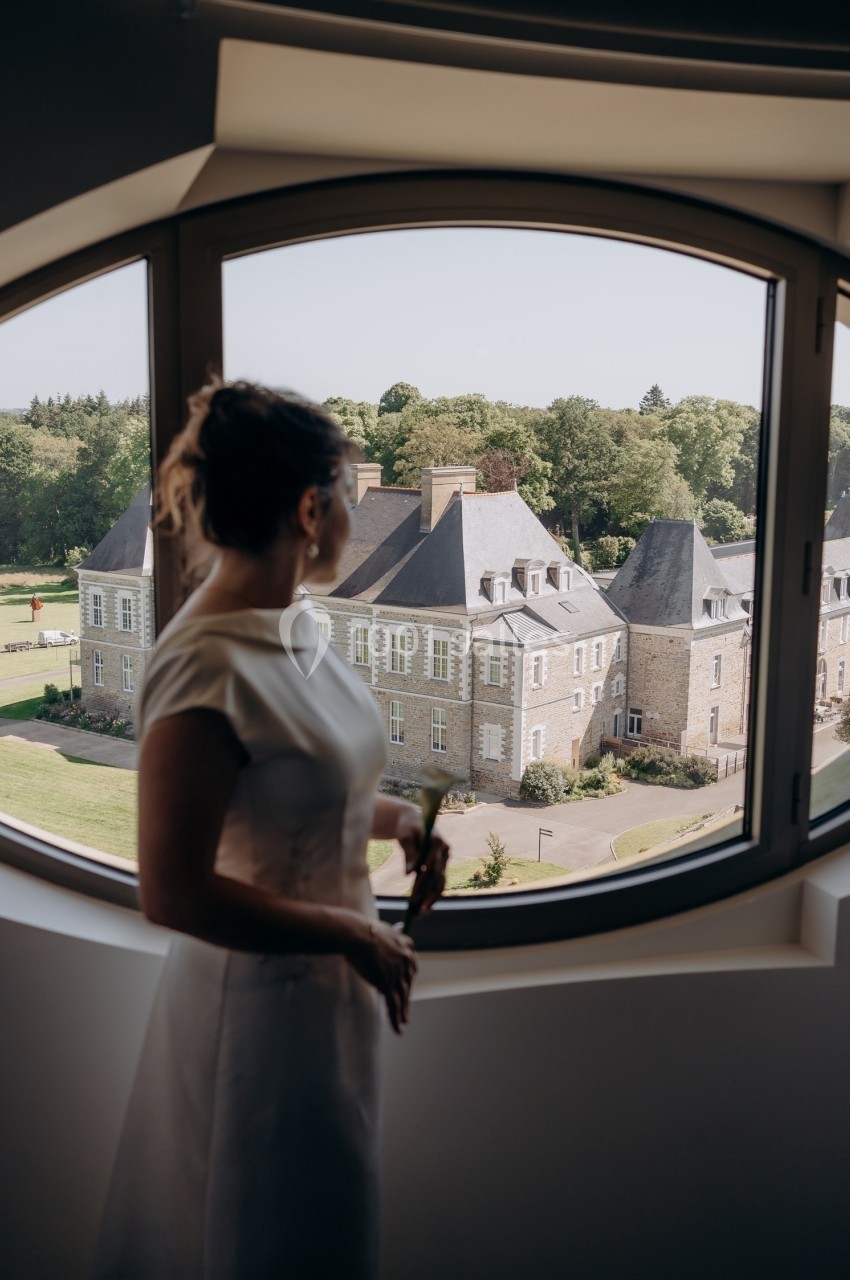 Une femme en robe blanche regarde par une fenêtre ronde un château entouré de verdure.
