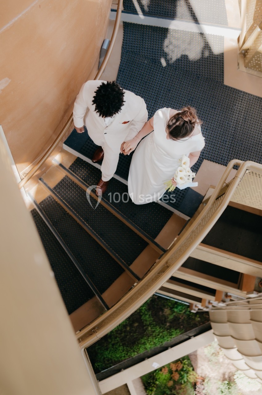 Un couple en tenue de mariage monte un escalier en colimaçon, vu de dessus.