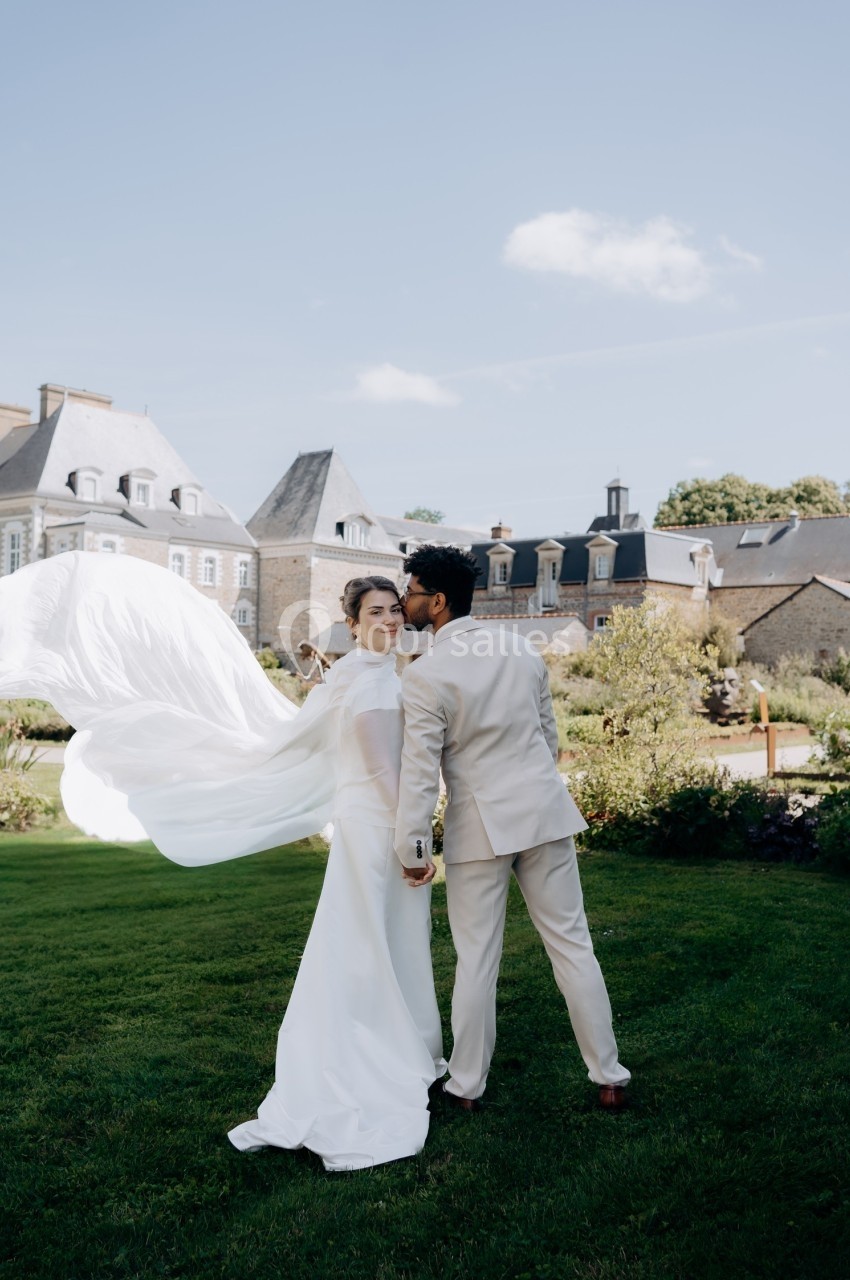 Un couple en tenue de mariage pose dans un jardin devant un bâtiment historique sous un ciel dégagé.