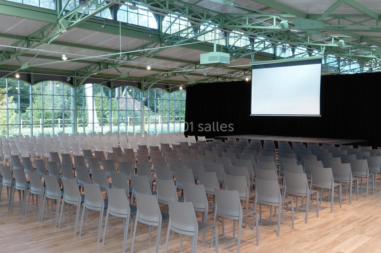 Salle de conférence lumineuse avec des rangées de chaises grises, une scène et un écran de projection.