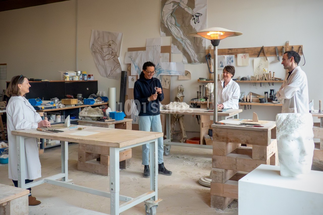 Des personnes travaillent dans un atelier d'art, entourées d'outils, de sculptures et de croquis accrochés au mur.