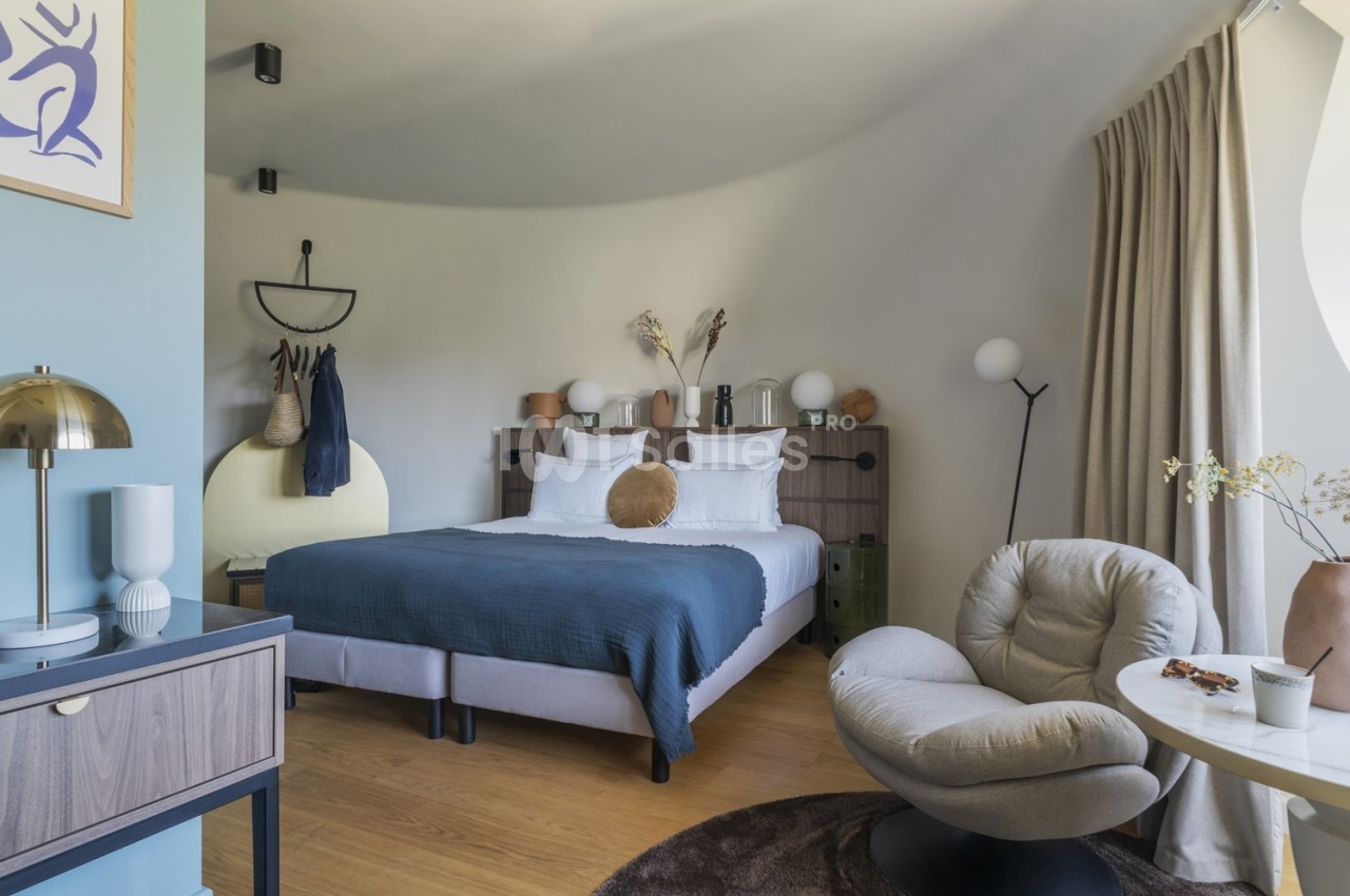 Chambre moderne avec lit double, fauteuil, bureau, décoration minimaliste et lumière naturelle filtrée par des rideaux.
