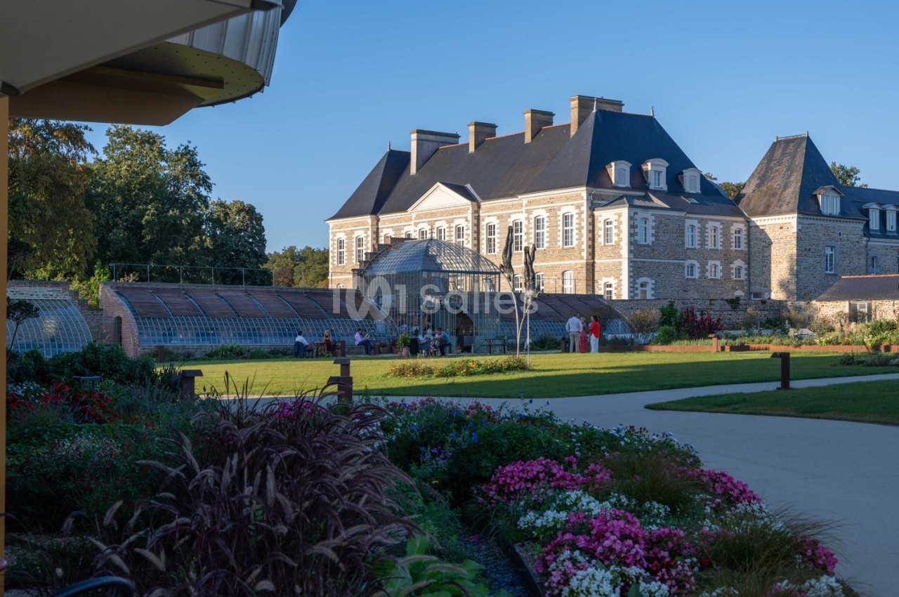 Jardin fleuri avec allée menant à un château en pierre et une serre, sous un ciel dégagé en fin de journée.
