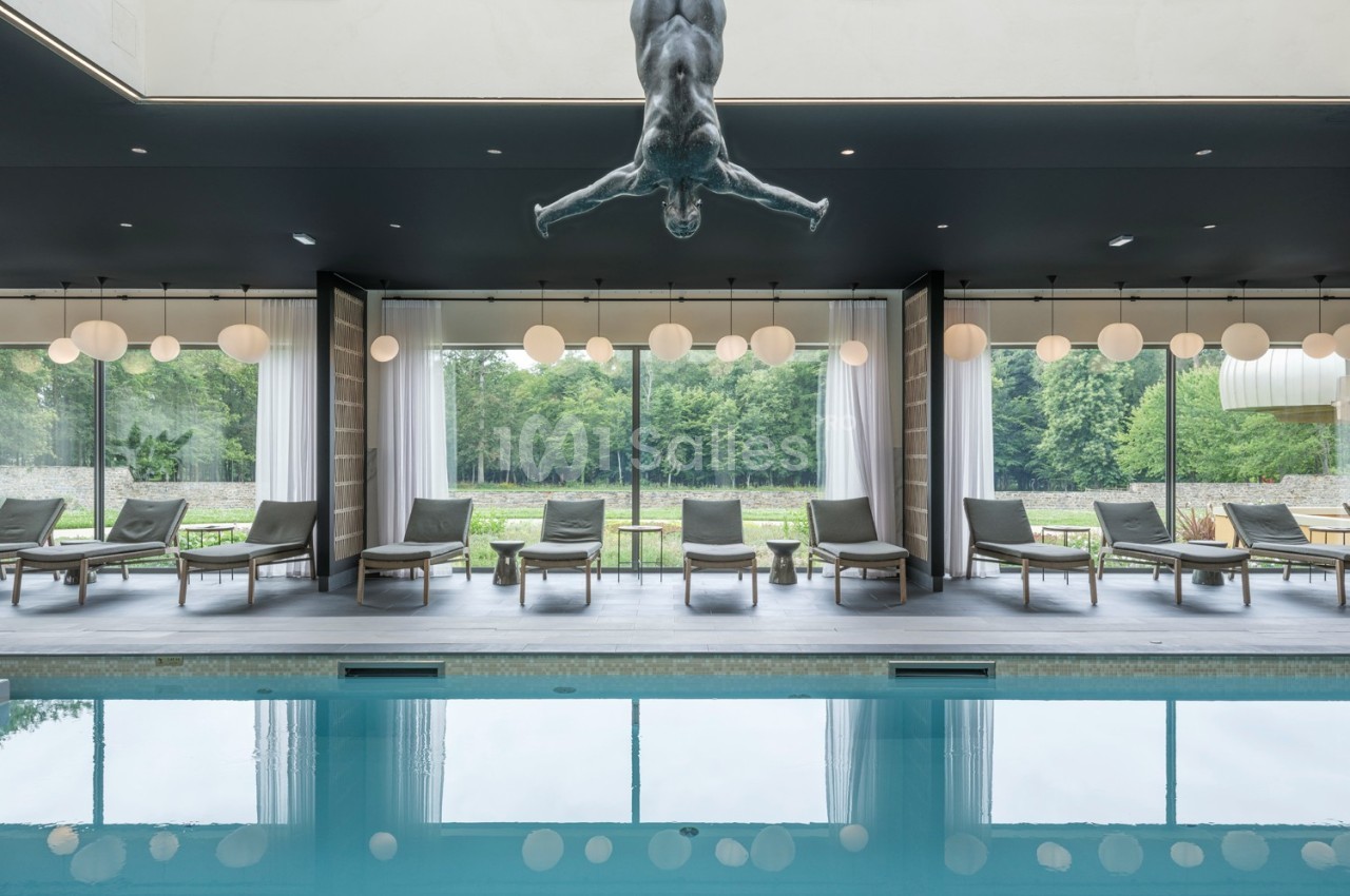 Un bassin intérieur avec des chaises longues alignées devant de grandes baies vitrées donnant sur un jardin.