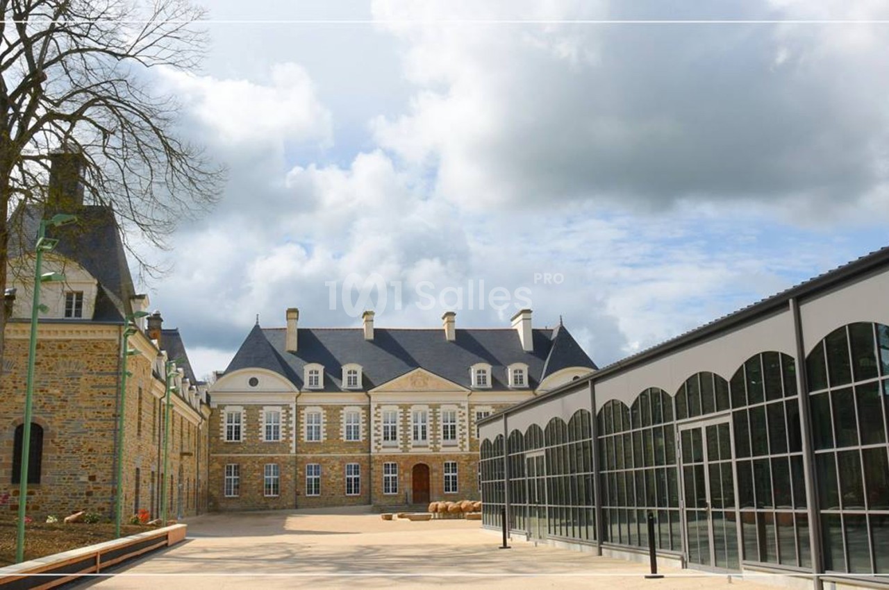 Façade d'un château en pierre avec toit en ardoise, à côté d'une structure moderne en verre sous un ciel nuageux.