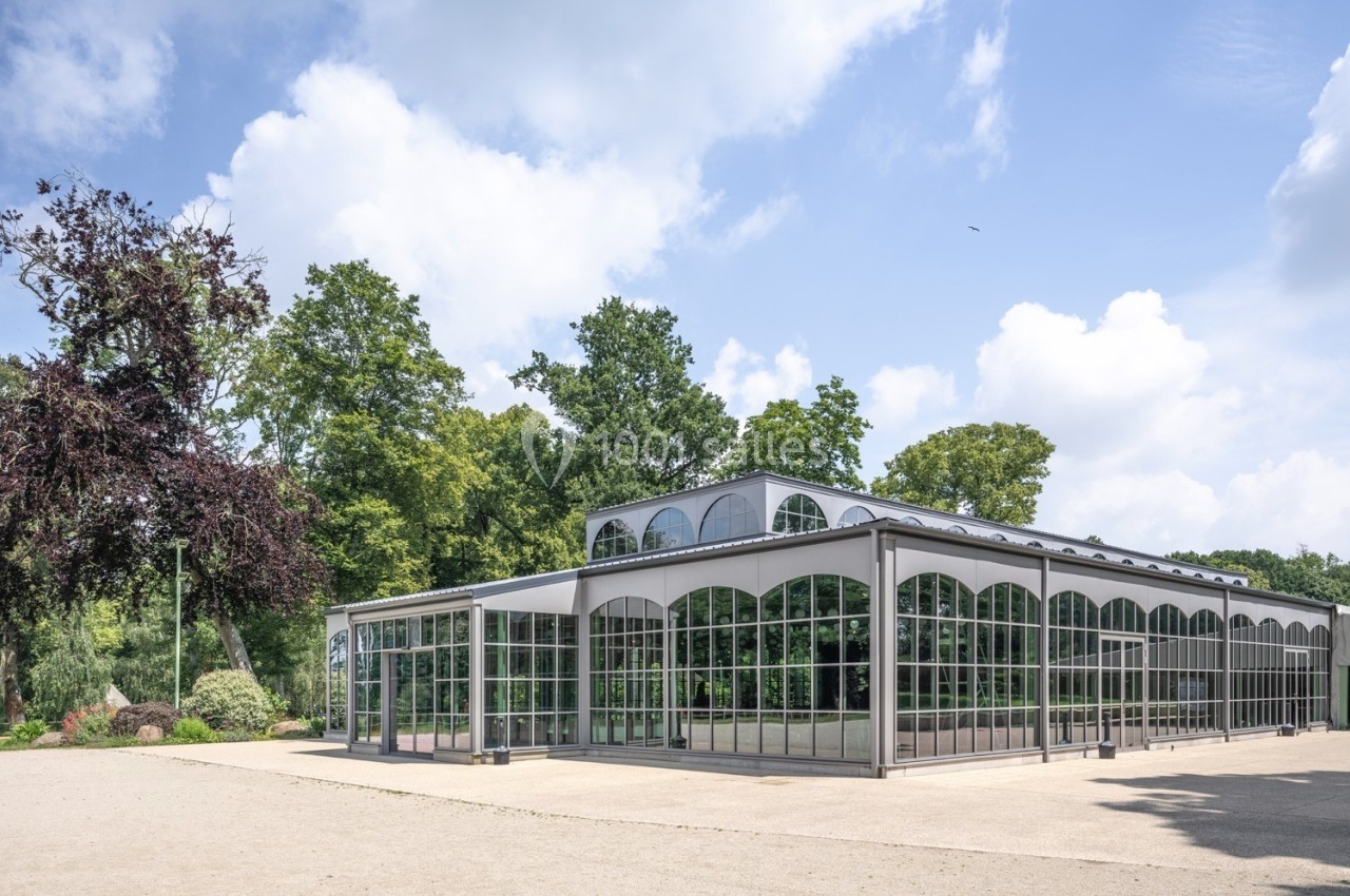 Serre moderne en verre et métal entourée d'arbres et d'un ciel partiellement nuageux.
