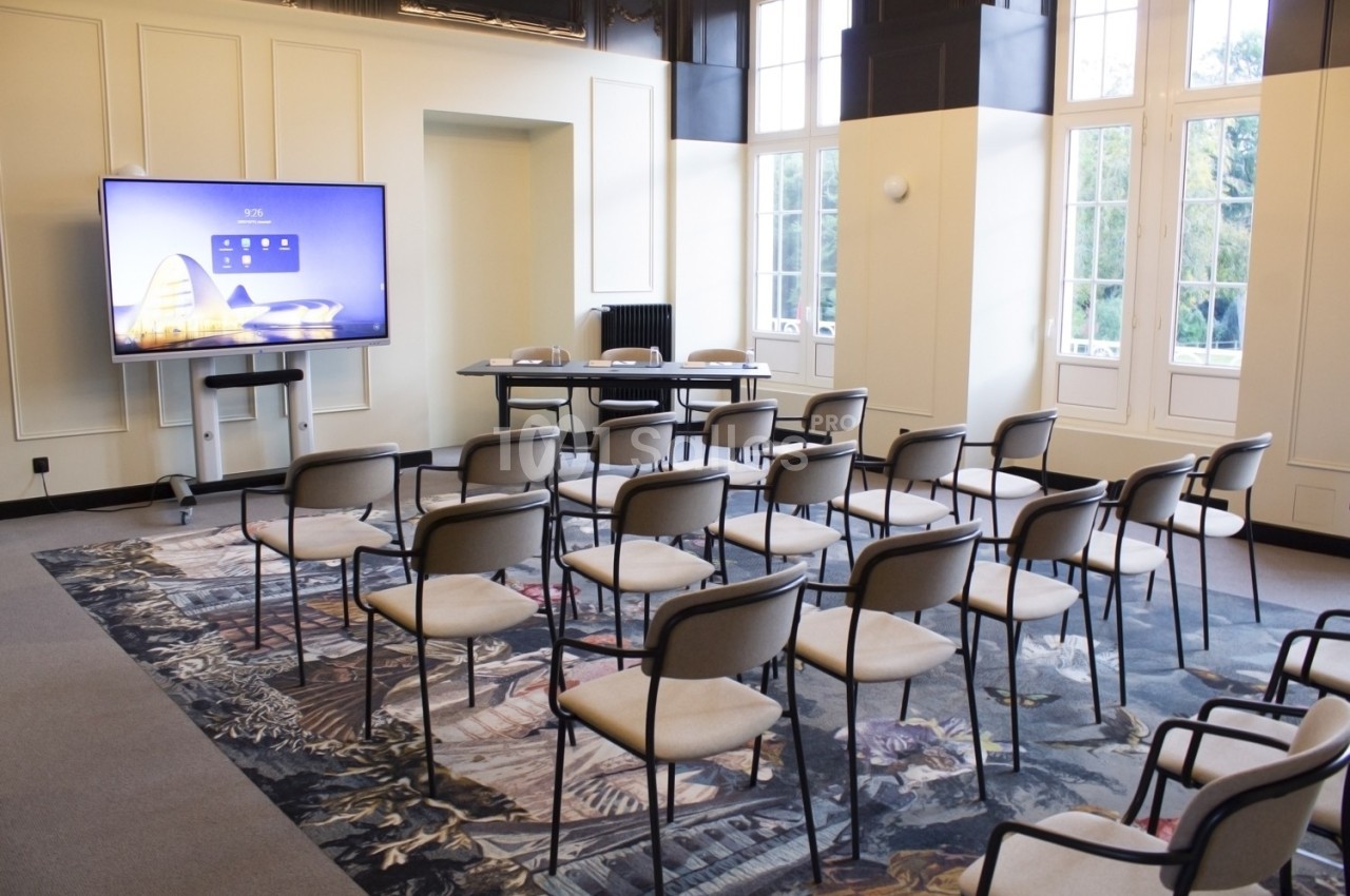 Salle de réunion lumineuse avec rangées de chaises, écran interactif et grandes fenêtres donnant sur un extérieur verdoyant.