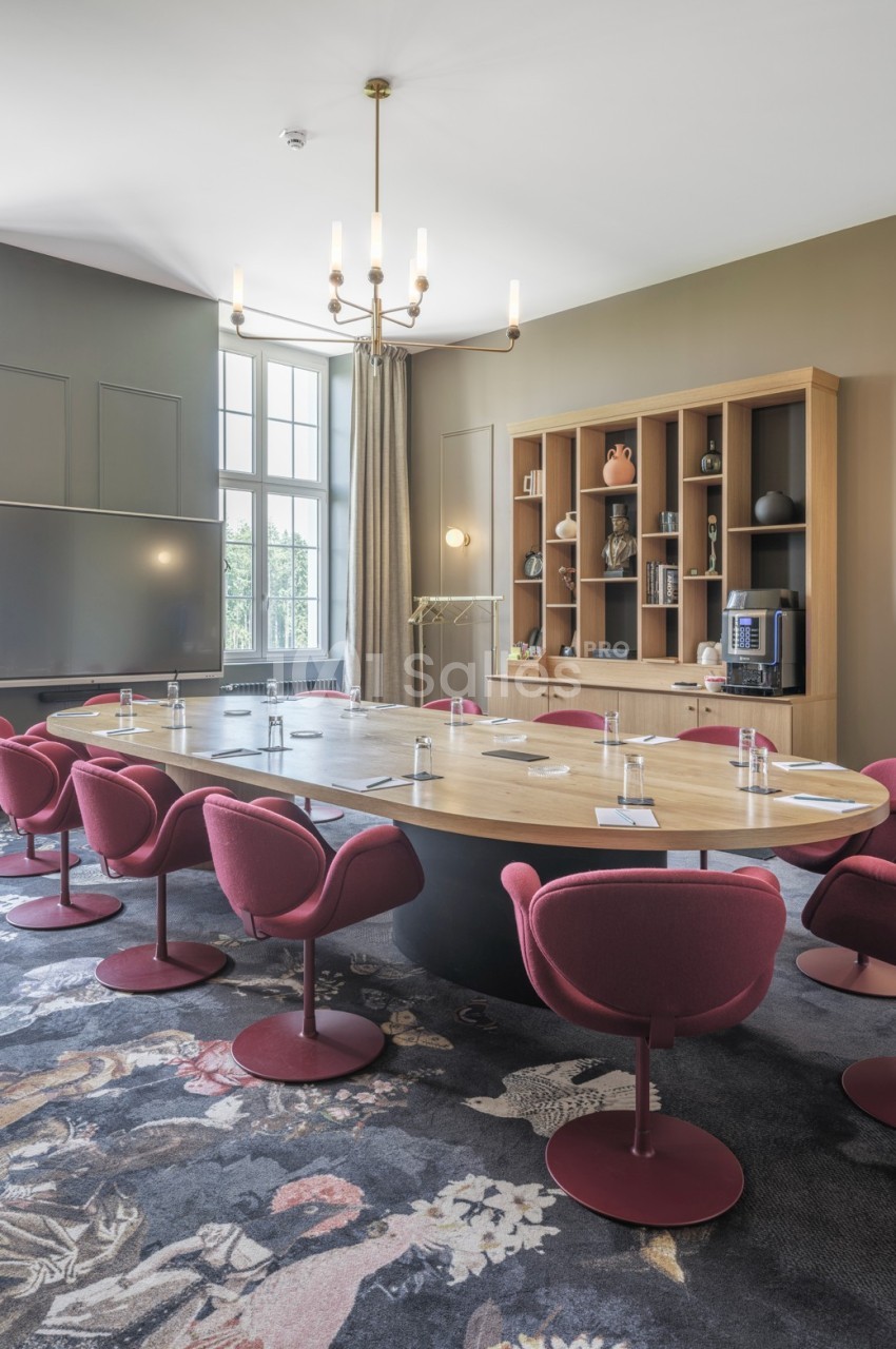 Salle de réunion lumineuse avec une grande table ovale en bois, chaises rouges, étagères et tableau blanc.