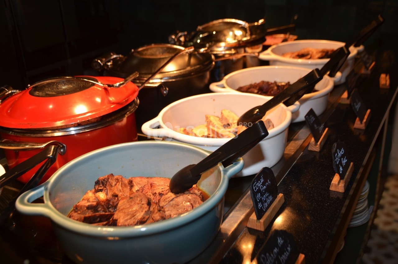 Buffet avec plusieurs plats chauds présentés dans des casseroles colorées, accompagnés de pinces de service.