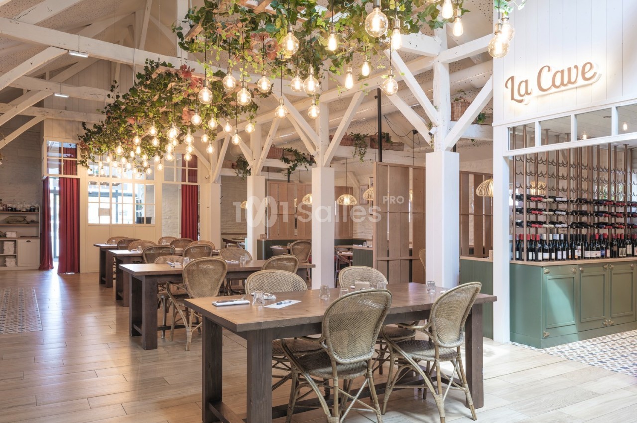 Salle de restaurant lumineuse avec tables en bois, chaises en rotin, décor végétal suspendu et espace cave à vin visible.