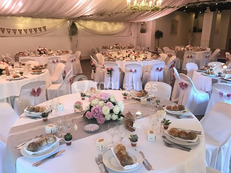 Salle de réception décorée pour un mariage, avec tables rondes dressées, nappes blanches et arrangements floraux.