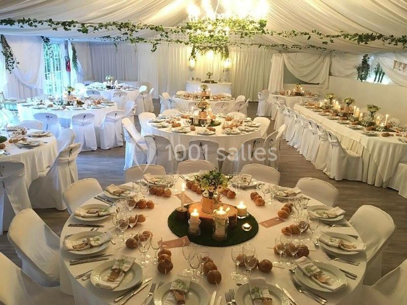 Salle de réception décorée avec des tables rondes et rectangulaires, nappes blanches, bougies et guirlandes végétales.