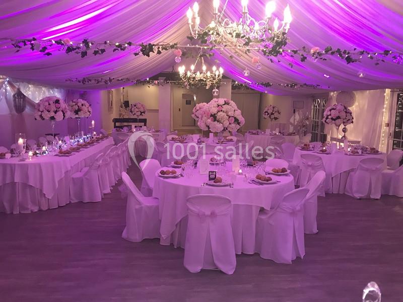 Salle de réception décorée avec des tables rondes, nappes blanches, fleurs roses et éclairage violet sous un plafond drapé.