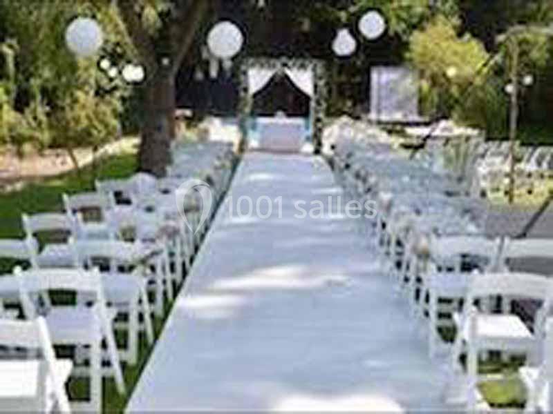 Allée centrale décorée pour une cérémonie en plein air, entourée de chaises blanches et de lanternes suspendues.