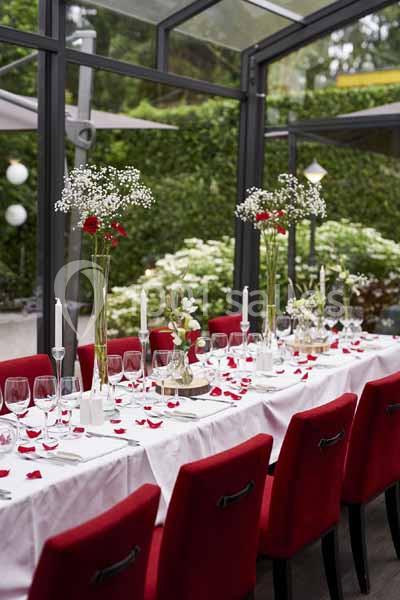 Table décorée avec nappes blanches, pétales rouges, chandeliers et vases fleuris, dans une salle vitrée donnant sur un…
