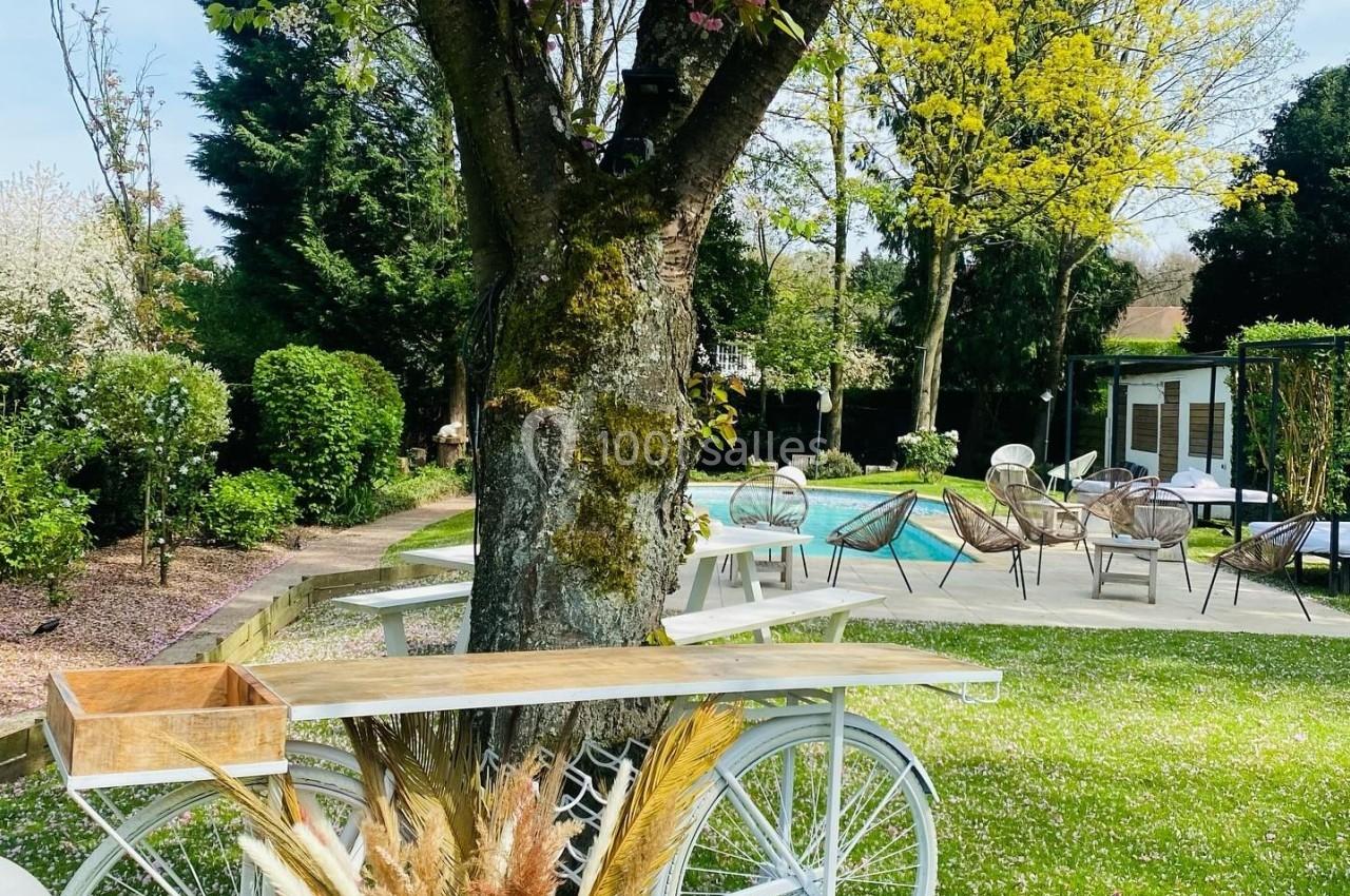 Un vélo blanc décoratif avec des fleurs séchées est placé sous un arbre dans un jardin verdoyant près d'une piscine.