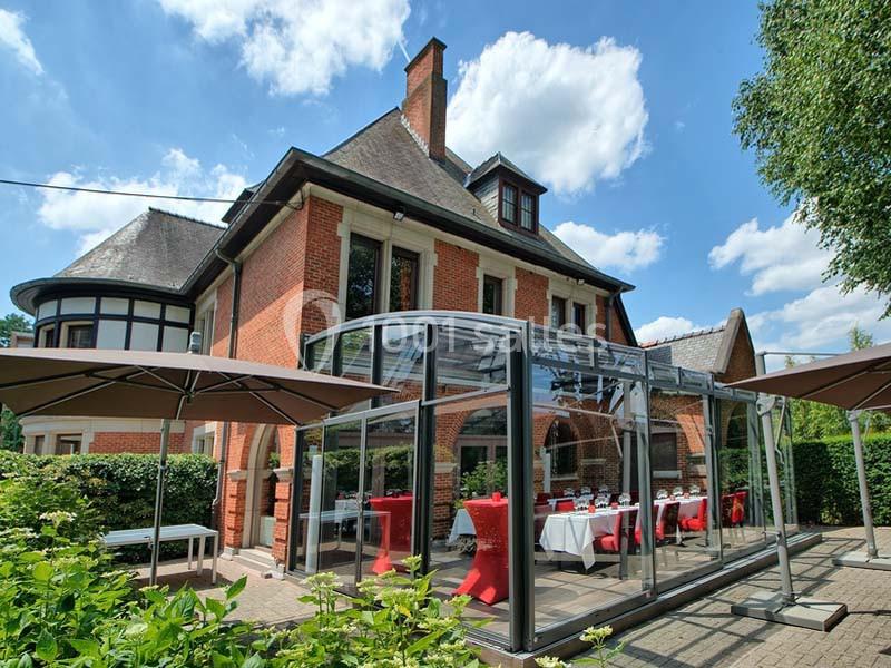 Façade d'une maison en briques rouges avec une terrasse couverte en verre et des parasols dans un jardin verdoyant.
