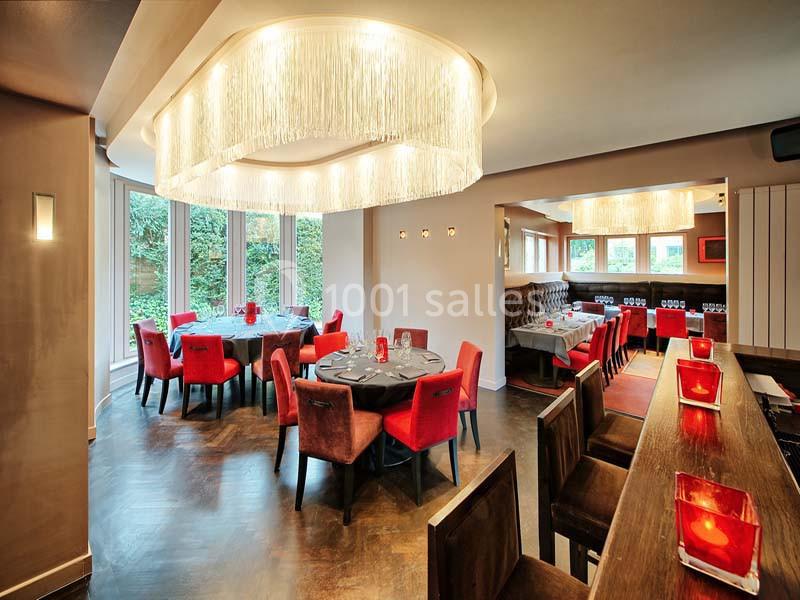 Salle de restaurant élégante avec tables rondes, chaises rouges, grandes baies vitrées et lustres modernes.