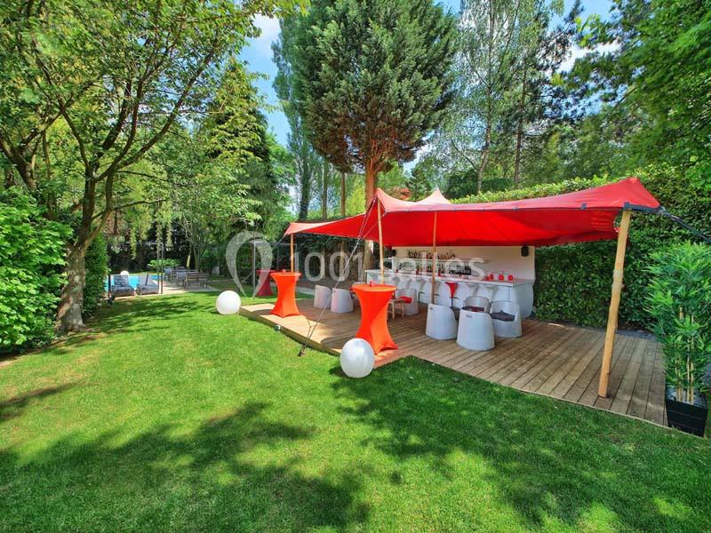 Terrasse extérieure avec tables et chaises sous une tente rouge, entourée de verdure et d'arbres.