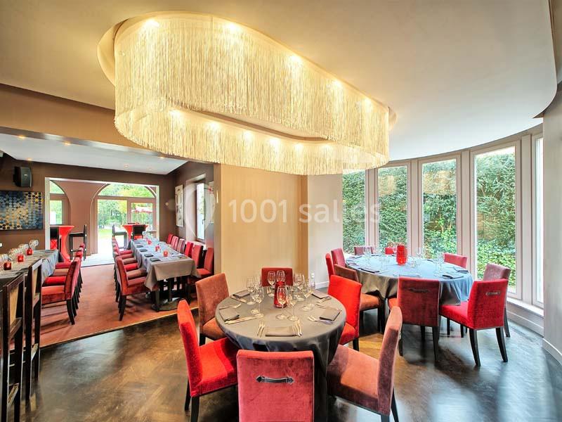 Salle de restaurant lumineuse avec grandes baies vitrées, tables dressées et chaises rouges, éclairée par un lustre moderne.