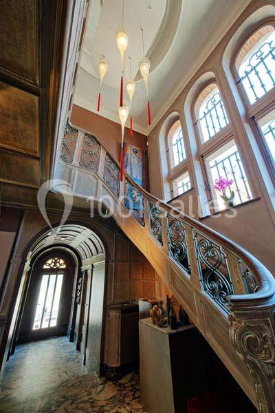 Escalier en bois sculpté avec rampe en métal, lustres suspendus et grandes fenêtres laissant entrer la lumière naturelle.